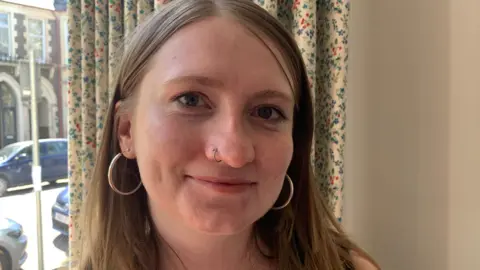 BBC Marianne looks at the camera, she's wearing hoop earrings and has a nose piercing. She's smiling and a curtain and window overlooking the street is visible behind her. 