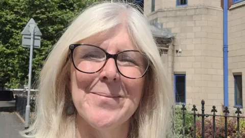 Allison Gardner An woman with long bright white hair and glasses smiling at the camera. She stands in front of a grey/brown brick building with the back of a road sign near to her. 