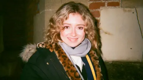 Getty Images Kate Shemirani, pictured in close-up with a long lens at an anti-vaccine protest in 2020. She wears a black dress with buttons at the shoulder and has long blonde hair that hangs in loose curls over her right shoulder. She is smiling brightly.