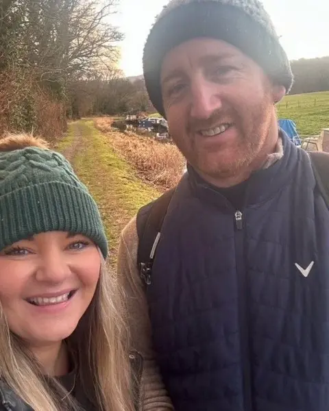Siobhan Gorman A selfie of Siobhan and her partner who are in the countryside with a canal behind them.  Both are smiling at the camera and wearing bobble hats. She has long wavy blonde hair and  is smiling. He has a cropped beard. 