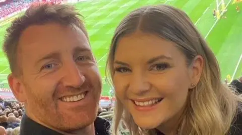 Siobhan Gorman A selfie of Siobhan and her partner who are in a  rugby stadium. Both are smiling at the camera from the seats with the pitch behind them. She has long wavy blonde hair and  is smiling. She is in an office. He has short ginger hair and a cropped beard. 