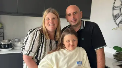 Jenni Whitmore Jenni with her husband Gareth and young daughter Carys in their kitchen. Jenni has her blonde hair down and is wearing a white, black and green dress. Her husband is wearing a black collared t-short. Her daughter is wearing a yellow hoodie and has long brown hair. 