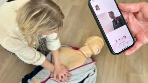 STEVE HUBBARD/BBC A woman attempts CPR on a sim dummy used to simulate emergency situations. The dummy is dressed in a zip up tracksuit top and has its head turned to one side. You can see its teeth. The woman has her arms out straight in front of her and has clasped one hand on top of the other on the dummy's chest. She has blonde hair and is looking at his chest to check for breaths. A phone is held to the right of the screen by a second person looking at a video call. It shows an advanced paramedic from the East of England Ambulance Service sat at a computer, guiding her on what to do. 