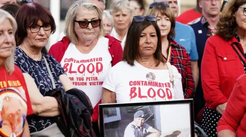 PA Media A group of family members protesting in May 2024 outside parliament. They are holding a photograph of their loved one and wearing white t-shirts saying "contaminated blood" in red writing.