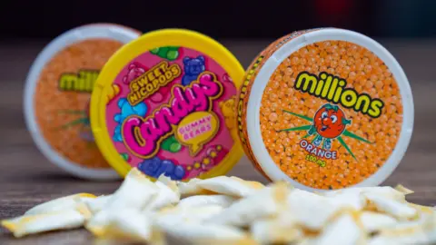 BBC A photo of three small tubs of nicotine pouches. One is orange and has a picture of a smiling orange on the cover with loads of small oranges under neath the word Millions. It does not mention the word nicotine. The Candys pack is pink with colourful gummy bears on it. It says "Sweet nicopods". 
