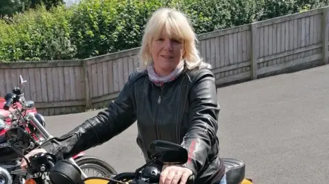 Mel Wallace Mel Wallace is a 59-year-old woman with shoulder-length blonde hair. She is sitting astride a yellow motorbike and is holding the handlebars. Mel is wearing a leather jacket and a light scarf around her neck. There are several motorbikes to her right and she seems to be in a car park with a wooden fence behind her, which is in front of a row of bushes