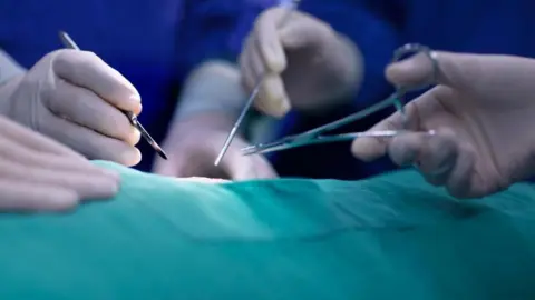 Getty Images Stock photo of three hands with white gloves on holding various medical tools as surgery is carried out. The patient is covered in a green gown, and the surgeons are in blue scrubs.