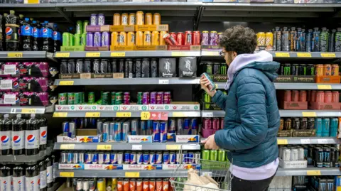 Getty Images A young man, dressed in a puffer coat, shops in a supermarket, picking up a can of energy drink and reading the nutritional information printed on the back