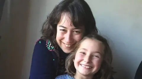 Merope Mills Photo of Martha Mills as a young girl with her mum, Merope behind her. Merope he has long brown hair, wears a dark grey top and is smiling at the camera. Martha has light brown/blonde hair and smiles broadly at the camera. She wears a light blue patterened outfit. 