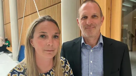BBC Sarah Hawkins, who has long blonde hair and is wearing a flowery top, stands to the left of Dr Jack Hawkins. He has stubble and is wearing a blue gingham shirt and dark suit jacket.