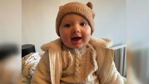 Family handout A baby boy is smiling widely. He is wearing a fluffy coat and knitted hat. He looks happy. 