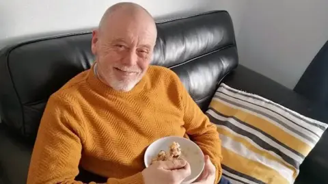 Family photo Ian Philip sits on a black leather sofa, smiling at the camera. He is wearing a mustard coloured jumper and midway through eating a bowl of cereal, which he is holding with one hand, a spoon in the other.
