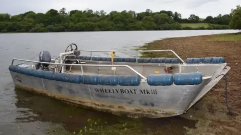MAIB The picture shows a small aluminium boat called a Wheelyboat Mk III, designed for wheelchair access. It has safety rails around the sides and blue cylindrical buoyancy fenders attached along the edges. The boat is equipped with a steering console and an outboard motor at the rear. It is positioned on the edge of a lake, partly on muddy ground, with water and trees in the background.
