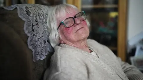 Nick Woolley/BBC Jean, an elderly person with white hair is seated in a cushioned chair with a patterned fabric cover. The person is wearing a light beige knitted sweater. In the background, there is a wooden cabinet with glass doors displaying various items.