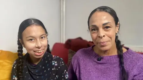 Phil Shepka/BBC Neave and Natasha are sitting indoors on a sofa. Both have long, dark hair styled in braids/pigtails. Natasha, who is taller, is wearing a purple knitted top with a small microphone clipped near the neckline. Naeve is dressed in a black top patterned with colourful stars and flowers. They are both looking at the camera and smiling. 