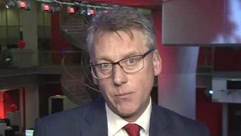 BBC Sir Julian Hartley looking into the camera. He wears glasses, a blue suit jacket, white shirt and red tie
