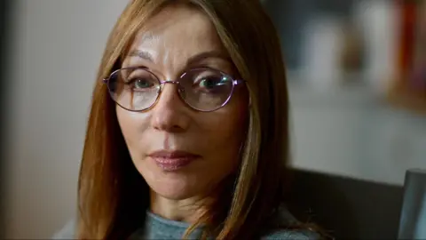 JAMIE NIBLOCK/BBC Sharon, Amy's mother looks at the camera lens. She is framed slightly to the left of the picture and has light brown to dark blonde, long, straight hair. She has chrome-framed rounded glasses on and is wearing light make-up. She has a light grey wool jumper on and the very top of her shoulders are in the frame.