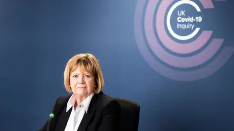 Piranha Photography Baroness Heather Hallett white shirt and a back jacket while sitting at a desk in front of the UK Covid-19 inquiry logo, which is made up of several concentric pink circles.