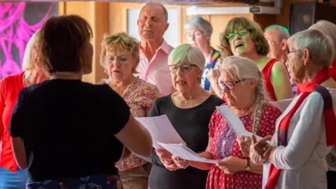 Sounds Better CIC A group of people singing together. They are all holding a few papers and looking down at them whilst singing. There is a person with their back to the camera leading the group.