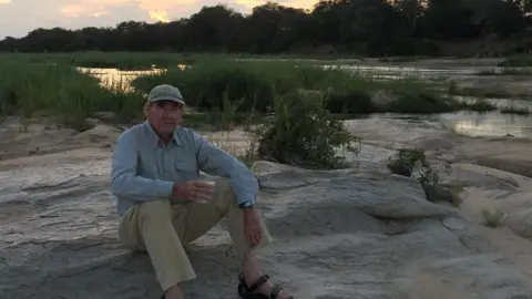 English family Sir Terence sits on a rocky area in light beige trousers and a blue shirt. Behind him is an orange and purple sunset. 