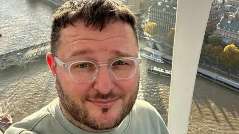 Mark Lewis This is a selfie of Mark Lewis, taken against the backdrop of Westminster, in London. He's a white man, in his 40s, and wears a grey sweatshirt with transparent round glasses. He has dark hair and dark facial hair too.