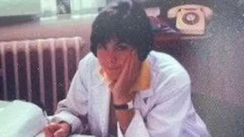 Dr Olwen Williams In this photograph from the 80s, Dr Olwen Williams is pictured at a desk. She's wearing a white overcoat with a yellow shirt underneath. She has a short black bob with a fringe. 