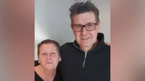 BBC A man and woman standing side by side with their arms around each other's shoulders. The woman has brown hair, tied back. The man has short grey hair. He is wearing glasses and a black jumper. 