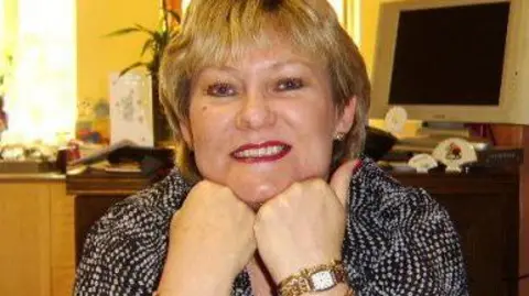 Other Colette Houliha, a woman with short blonde hair, wearing a black and white top - she has her chin rested on her hands and is smiling, she is inside with a computer monitor behind her 