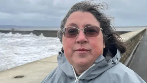 BBC Dawn Lord has light brown sunglasses on with her hair being blown back in the wind. She is wearing a light grey coat with the sea splashing against a sea wall in the background.