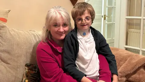 BBC A boy with a genetic condition is wearing small black-framed glasses, a white polo-shirt and a black fleece. He is smiling at the camera while sitting on the lap of his mum who is wearing a pink jacket. She has grey hair and is also smiling. They are both sitting in a lounge on a beige sofa which has a light-brown fluffy blanket.