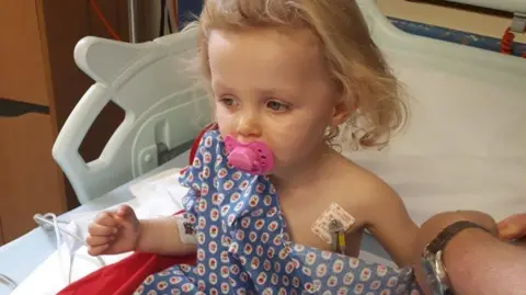 Nye family Young girl in a hospital bed wearing a blue bed-shirt. She looks poorly and has a pink dummy in her mouth. 