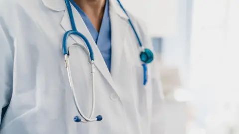 Rawlstock/Getty Images An unidentifiable doctor wearing a white jacket over a blue top. They have a stethoscope around their neck. 
