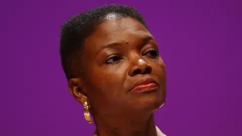 Baroness Amos, wearing gold earrings, look away from the camera before a purple background.