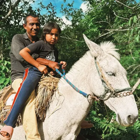 Ana's dad is smiling as he and Ana ride a donkey/horse - white with a straw saddle. Ana is wearing navy joggers and a black Adidas t-shirt, and her dad is wearing a dark shirt and yellow trousers   