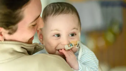 PA Media A young baby with dark hair looks past the camera, smiling. He has a feeding tube inserted into his nose and is chewing on of his hands.
A dark haired woman is holding him and she is pictured from behind.