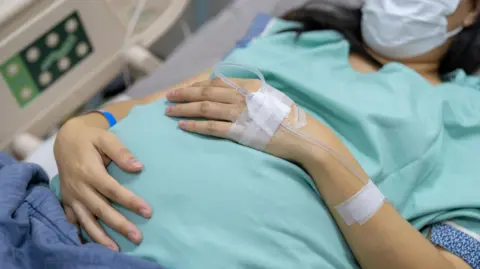 Getty Images A pregnant woman waits to give birth lying in a hospital bed, wearing a blue gown and mask, with a tube attached to the back of her hand