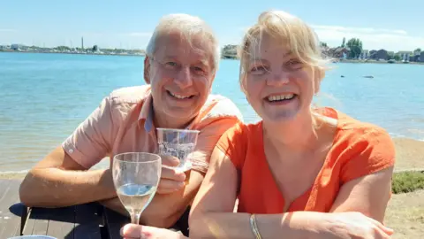 Family photo Chris and Ruth Stone-Houghton sit on a picnic bench, smiling into the camera. They are holding alcoholic drinks, and behind them is a body of water