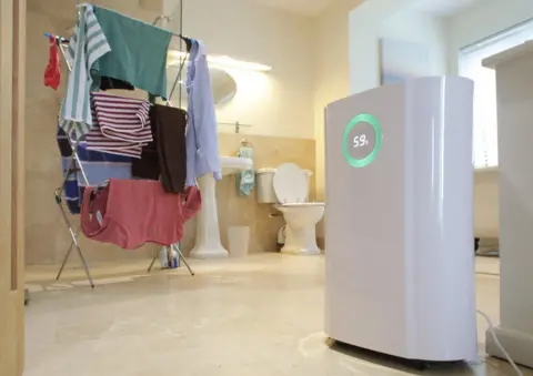 Getty Images A dehumidifier runs next to a pile of washing which is drying on a clothes rack in a large bathroom. The dehumidifier display reads 59%.