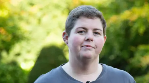 Abbie Harvey is stood in the gardens of the Parkland Place Rehabilitation Centre with green trees and bushes behind her. She's wearing a grey t-shirt and is looking directly at the camera.