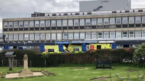 David Robinson / Geograph A long, wide building with numerous windows stands in the centre, with ambulances outside an entrance, a sign reads 'Darlington Memorial Hospital'. A park sits in front. 