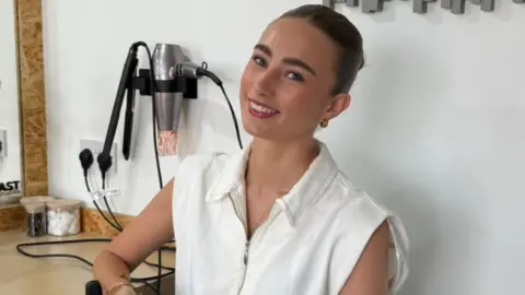 Vicky Evans A woman in a white top with her hair tied up smiles at the camera. Behind her are hair straighteners and a hair dryer