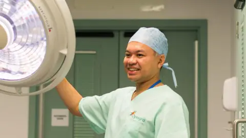 PA Media A man wearing medical scrubs in pale green, looking towards a large operating light.