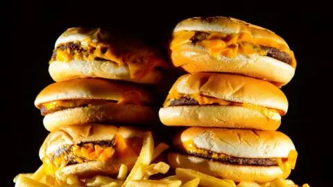 PA Media A stack of six cheeseburgers is piled on top of a portion of chips against a black backdrop. 