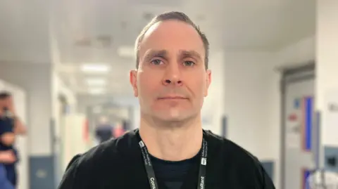 Dr Carl is looking directly at the camera. He has short brown hair and is wearing a black t-shirt. He has a black and white lanyard around his neck.