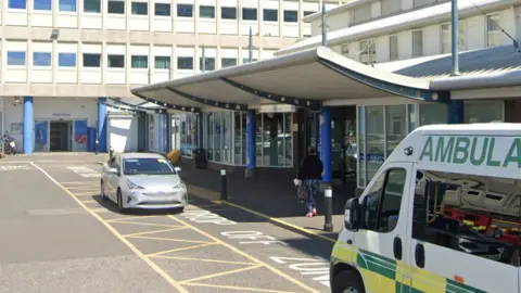 Google The outside of  North Tees Hospital An ambulance is waiting in the yellow hatched drop off zone and a silver car is also parked. Parts of larger hospital building can be seen in the background.