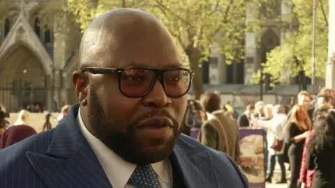 Lanre Haastrup is standing in front of crowds outsdie the Supreme Court. He is bald with a black beard. His is wearing black glasses and a blue suit. 