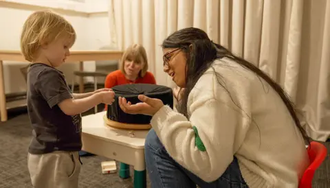 Kevin Church/BBC News A psychology researcher engages in a game with a young toddler. The researcher, on the right of the image, sits in a very small, plastic chair, so that she is at the correct height to engage with the young boy. The boy, about two years old, is looking at a sticker he just found, which was the aim of the game he is playing. 