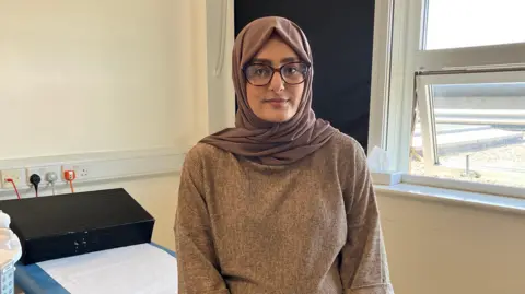 Komal is smiling at the camera, sitting on a bed in a doctor's office. She is wearing black glasses,  a brown hijab, brown top and blue jeans. 