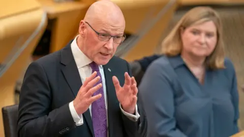 PA Media John Swinney, who has a bald head and glasses, speaks in the Scottish Parliament chamber. He is wearing a dark suit, white shirt and purple tie. 